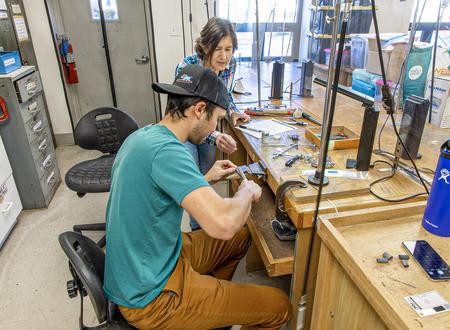 A man is seated working at a jeweler bench and a woman stands over him to the side guiding him