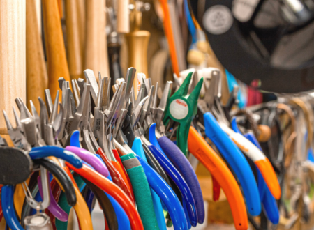 Jewelry pliers with different color handles are lined up and hanging on a rod and a pair of sunglasses can be seen hanging behind them
