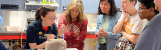 A Metalwerx instructor wearing a black shirt and apron demonstrates torch techniques to a group of students