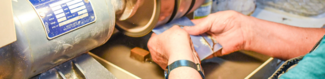 Two white hands hold a piece of square enamel art up to the wheel of Metalwerx Studio's lapidary grinder